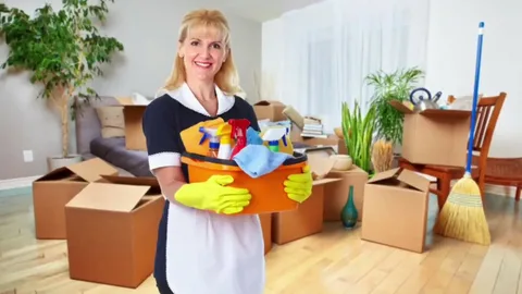 A smiling professional cleaner, dressed in a black and white uniform with yellow gloves, is holding a caddy full of cleaning supplies in a messy room filled with cardboard moving boxes.