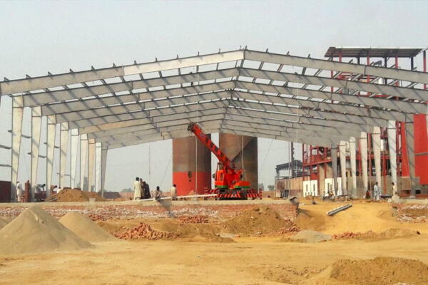 A Complete Guide to Industrial Warehouse Construction This image displays a Pre-Engineered Building (PEB) structure in its early construction phase, highlighting the erection of a steel frame for a large-scale facility. A crane and construction workers are actively involved on the site, demonstrating the process of assembling the prefabricated steel components.