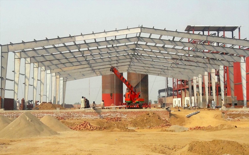This image displays a Pre-Engineered Building (PEB) structure in its early construction phase, highlighting the erection of a steel frame for a large-scale facility. A crane and construction workers are actively involved on the site, demonstrating the process of assembling the prefabricated steel components.